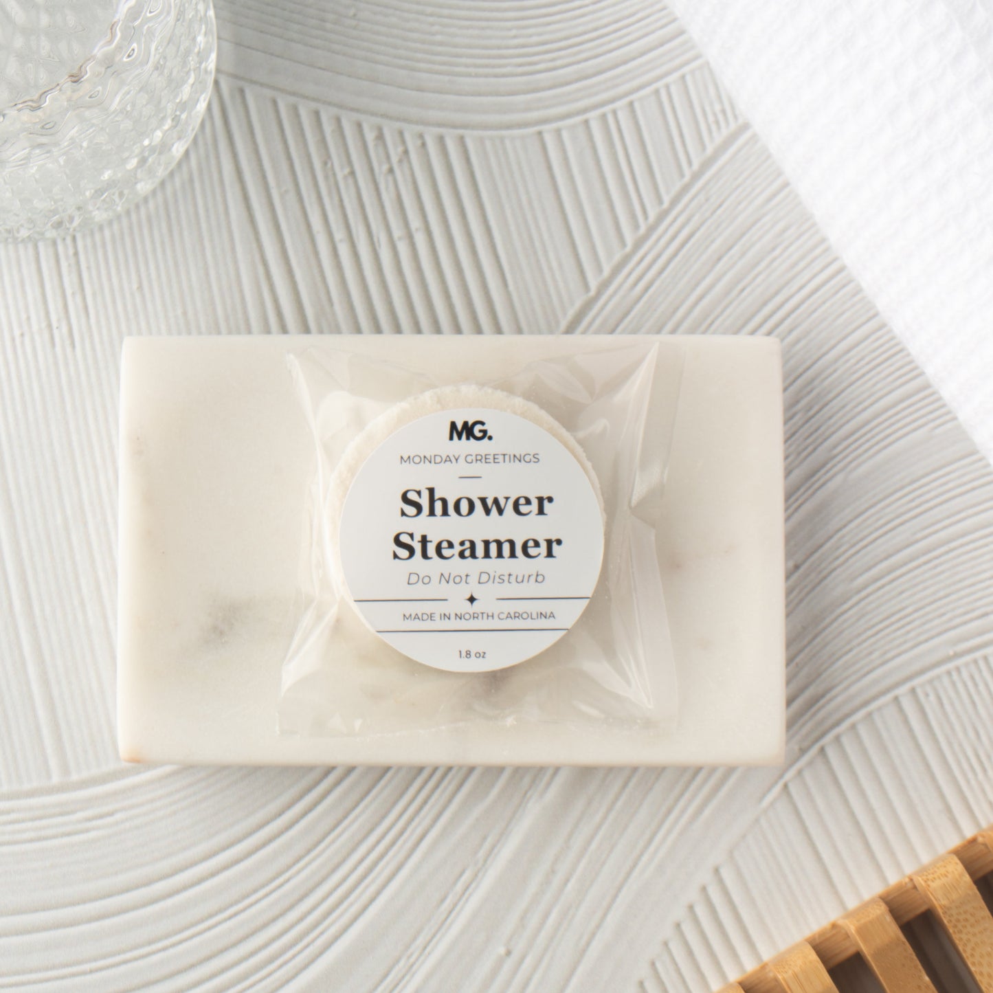 A packaged shower steamer from MG Monday Greetings placed on a textured white surface with a glass bowl and wooden slats as styled props.