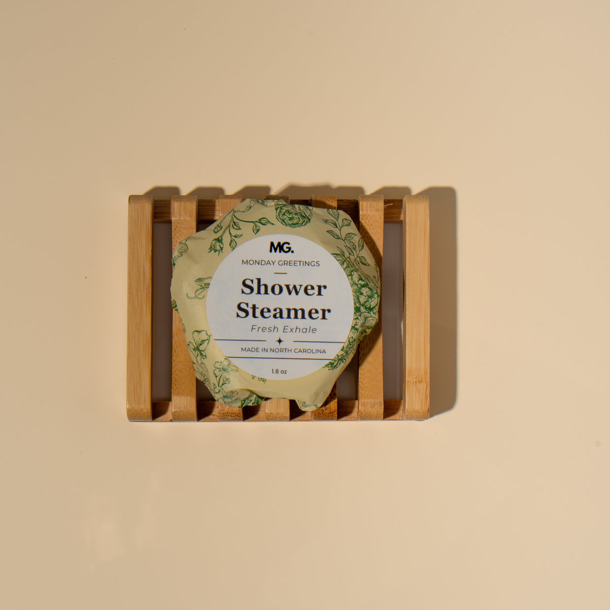 A round shower steamer wrapped in a green botanical-patterned paper sleeve, displayed on a small wooden slatted stand against a warm beige background. The label reads 'MG Monday Greetings Shower Steamer