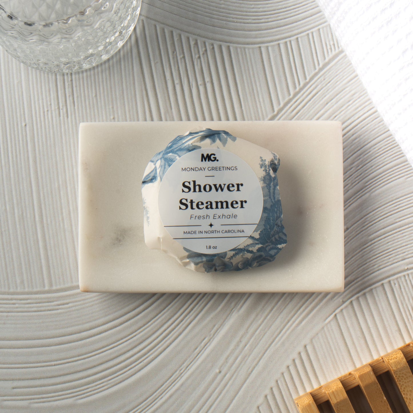 A single shower steamer packet wrapped in white paper with blue floral artwork sits on a rectangular white marble tray. The scene is styled on a textured light gray surface with a glass vessel at the top left and a wooden slatted object at the bottom right.