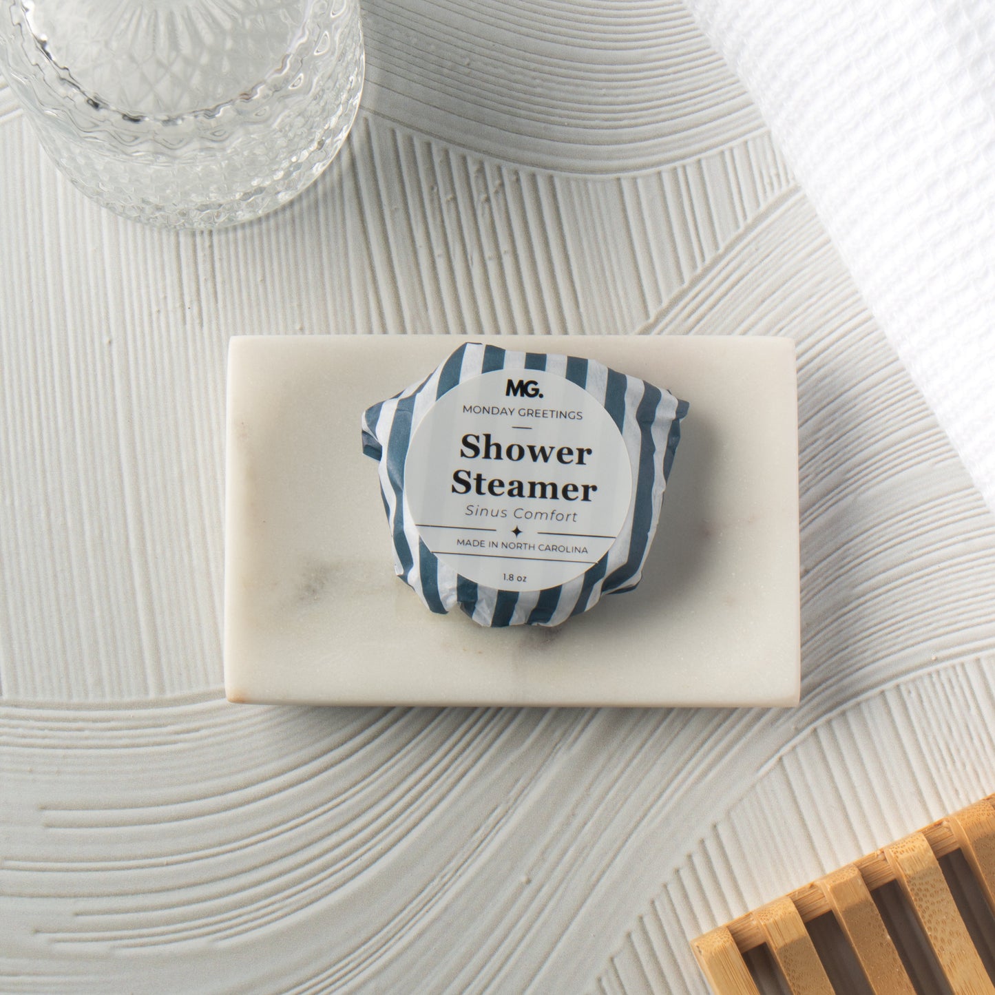 A wrapped shower steamer from MG Home Goods resting on a white marble soap dish, styled on a textured white surface with a glass jar and a wooden dish in the background.
