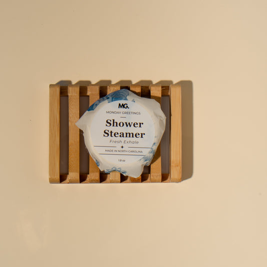 A packaged shower steamer sits on a small wooden slatted holder against a plain warm beige background. The package is white with blue watercolor-style edges and visible text including 'MG' and 'Shower Steamer'.