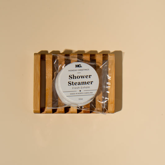 A packaged shower steamer by Monday Greetings, resting on a wooden slatted tray against a solid beige background.