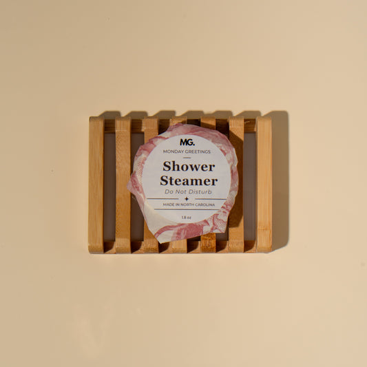 A packaged shower steamer resting on a small wooden slatted stand against a plain warm beige background. The round package has a pink floral border and reads 'M.
