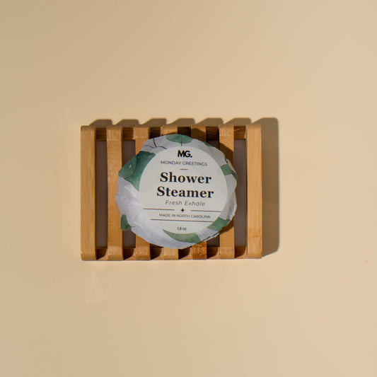 A round shower steamer wrapped in a white paper label with green accents, displayed inside a small wooden slatted box on a solid beige background. The label reads 'MG Monday Greetings Shower Steamer'.