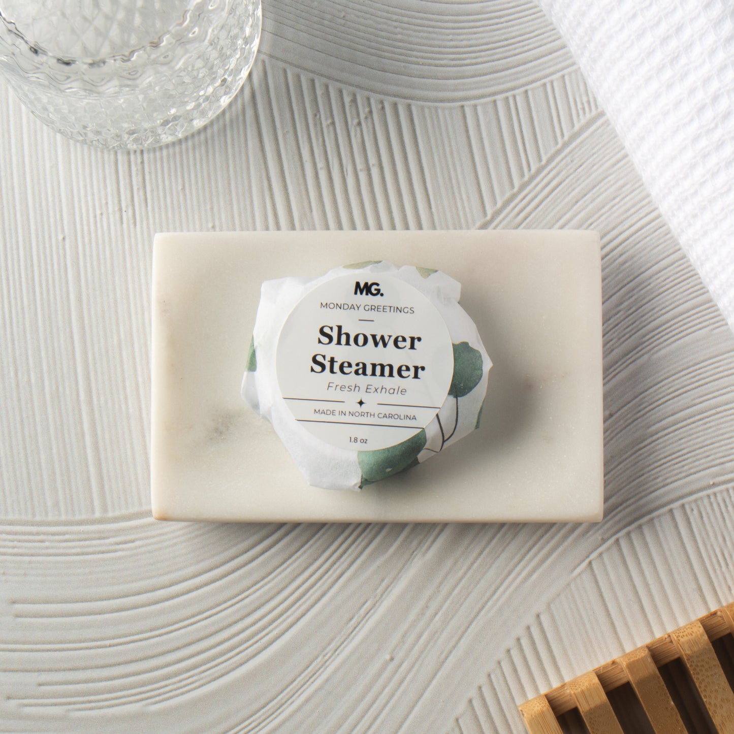A wrapped shower steamer from Morning Greetings rests on a rectangular cream soap bar, styled on a textured white surface with a glass jar, white towel, and wooden slats nearby.
