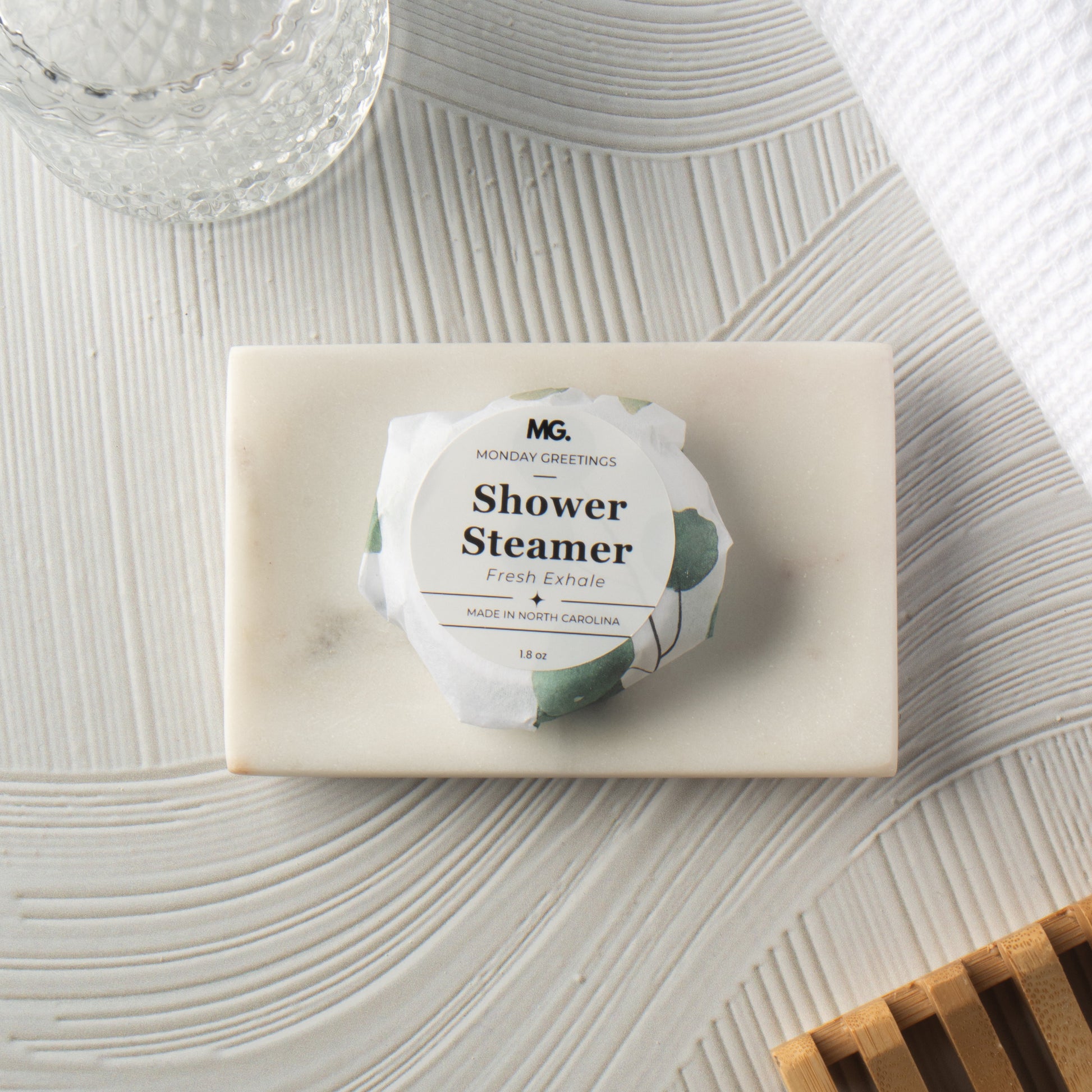 A wrapped shower steamer from Morning Greetings rests on a rectangular cream soap bar, styled on a textured white surface with a glass jar, white towel, and wooden slats nearby.