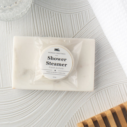 A packaged shower steamer bar from MG Monday Greetings, wrapped in clear plastic with a white circular label, placed on a textured white surface with a wooden slatted tray in the corner.