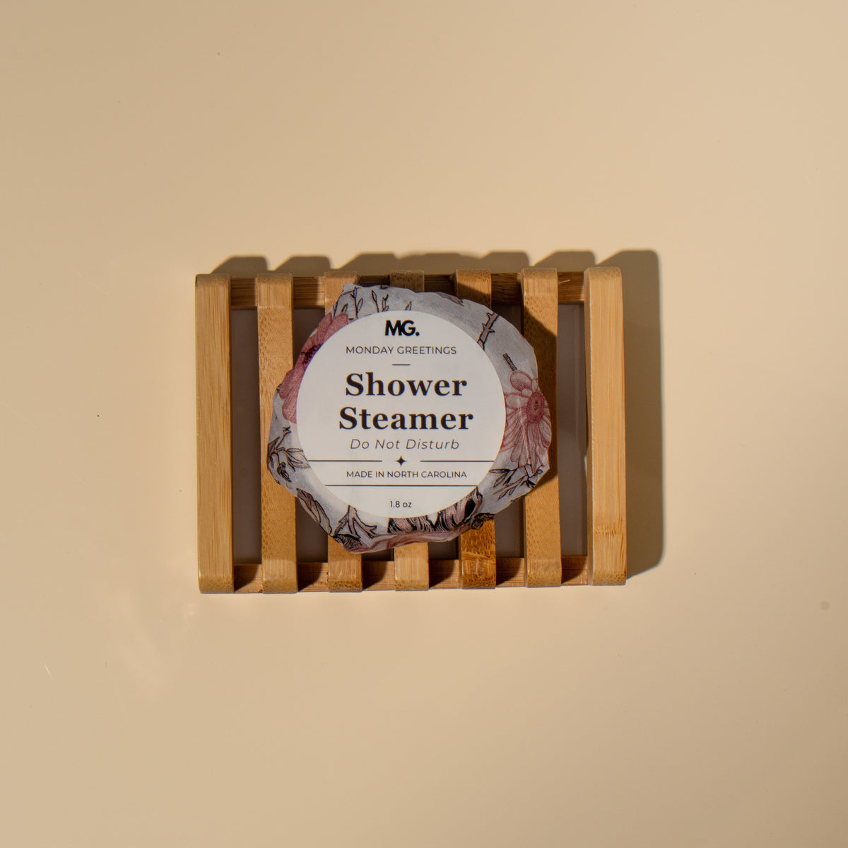 A single packaged shower steamer sits centered on a light wood slatted tray against a warm beige background. The round package has a floral label reading 'do not disturb 