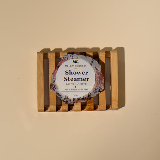A single packaged shower steamer sits centered on a light wood slatted tray against a warm beige background. The round package has a floral label reading 'do not disturb 