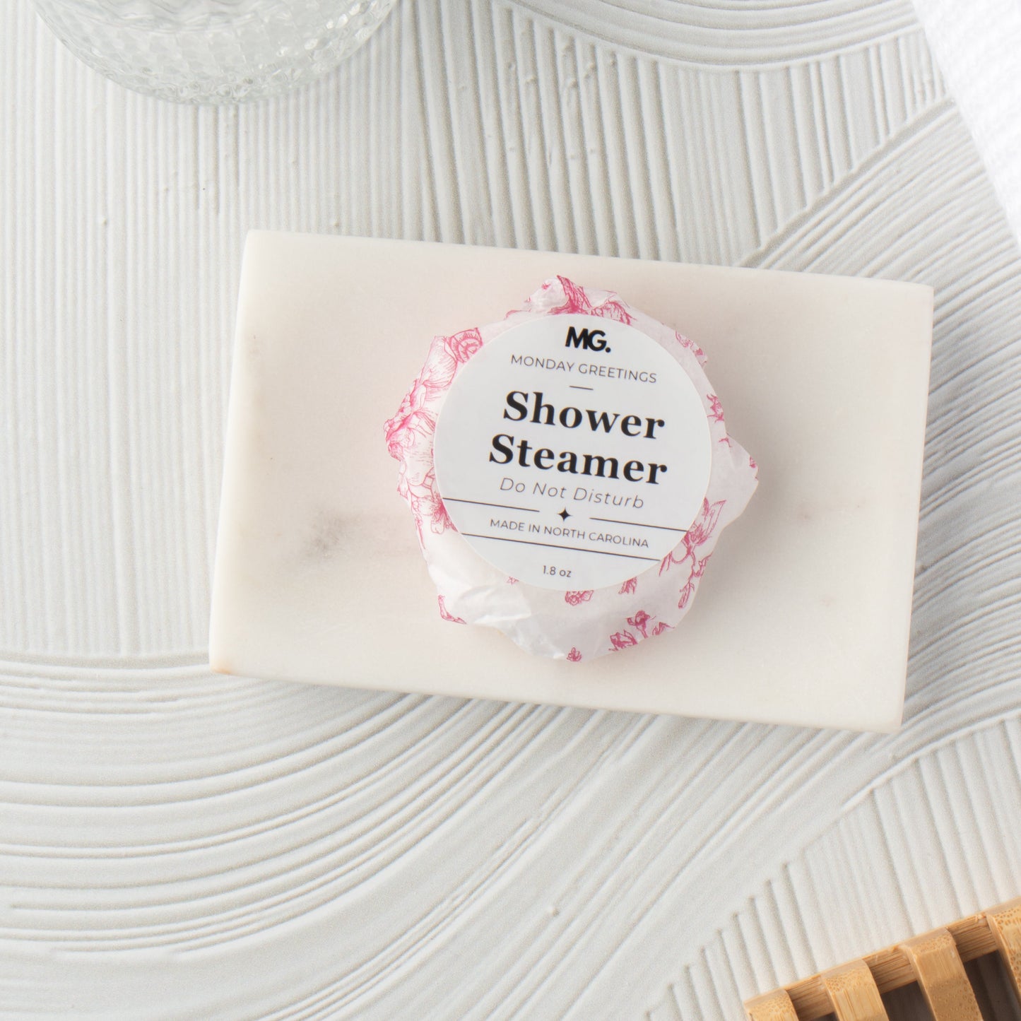 A wrapped shower steamer from Monday Greetings rests on a white marble soap dish, styled with a glass bowl and a wooden slatted coaster on a textured white surface.