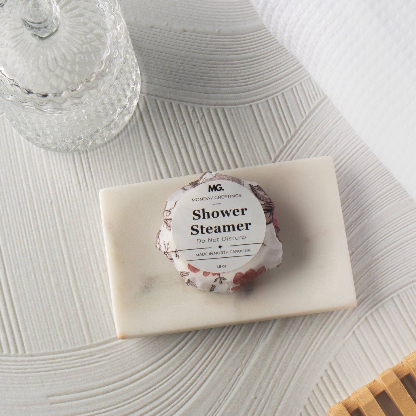 A wrapped shower steamer sits on a rectangular marble tray on a textured white surface, with a glass jar and white towel in the background.