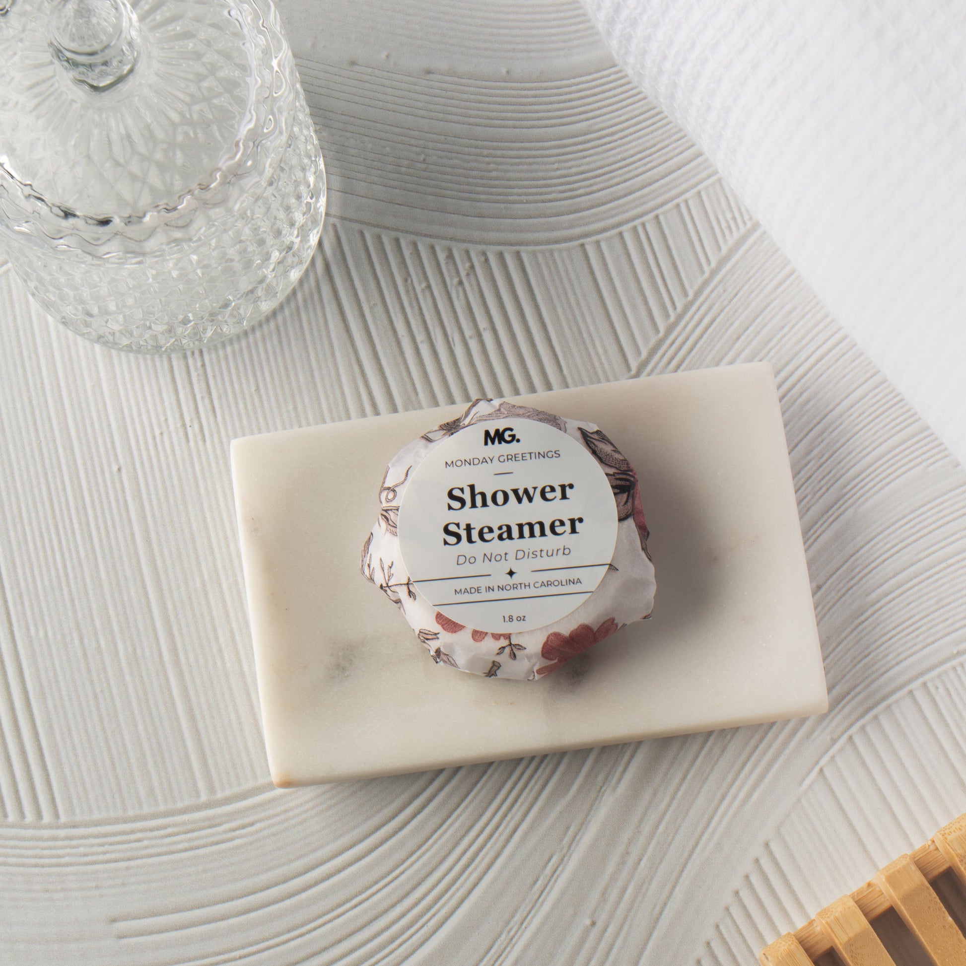 A wrapped shower steamer sits on a rectangular marble tray on a textured white surface, with a glass jar and white towel in the background.