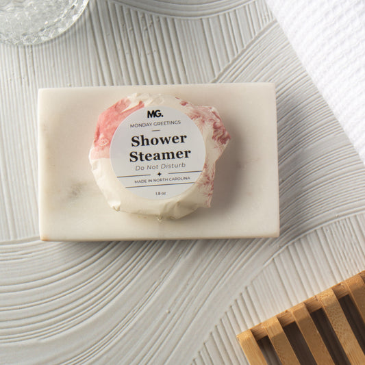 A wrapped shower steamer from Monday Greetings rests on a white marble soap dish, styled on a textured light gray surface with a wooden slatted tray in the corner.