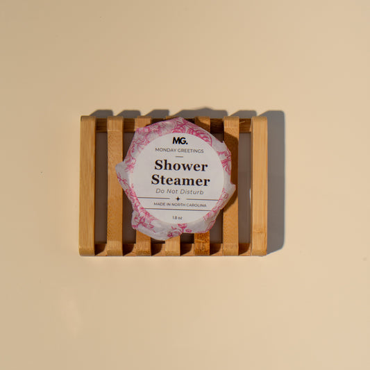 A round shower steamer wrapped in a pink patterned paper sleeve with a white center label reading 'MG  Shower Steamer displayed on a small wooden slatted stand against a warm beige background.