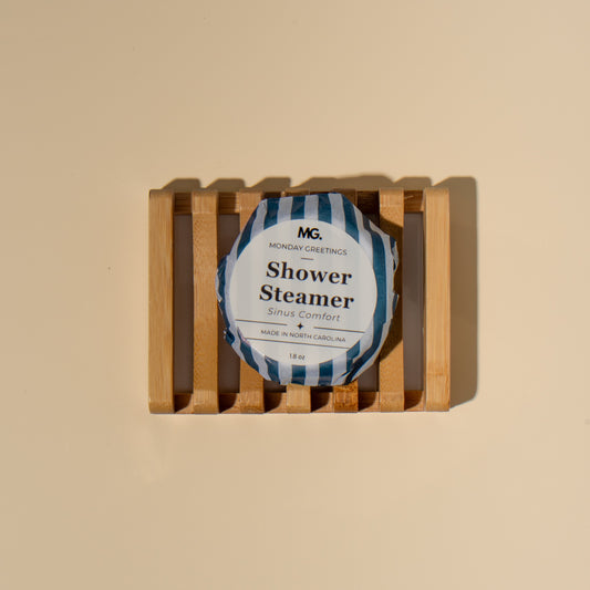 A single wrapped shower steamer sits centered on a small wooden slatted tray against a warm beige background. The product wrapper is white with blue diagonal stripes and black text reading 'MG' and 'Shower Steamer'.