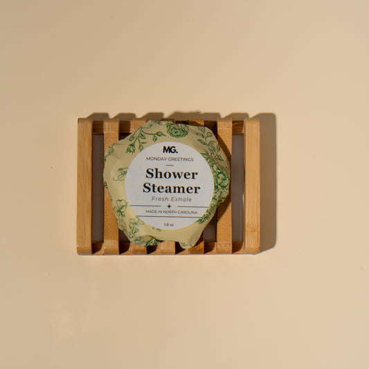 A round shower steamer wrapped in a green botanical-patterned paper sleeve, displayed on a small wooden slatted stand against a warm beige background. The label reads 'MG Monday Greetings Shower Steamer 